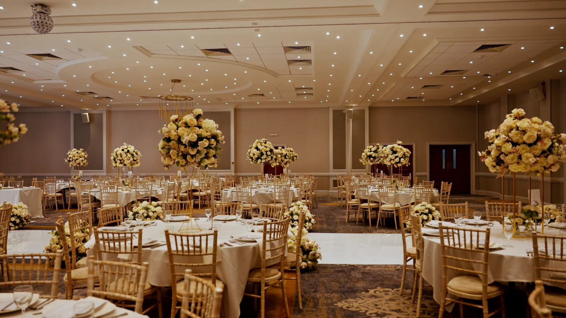An elegant hotel ballroom reception featuring round tables adorned with white linens, chiavari chairs, and towering floral centrepieces under a ceiling dotted with lights.
