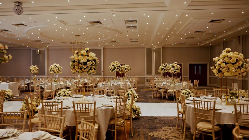 An elegant hotel ballroom reception featuring round tables adorned with white linens, chiavari chairs, and towering floral centrepieces under a ceiling dotted with lights.