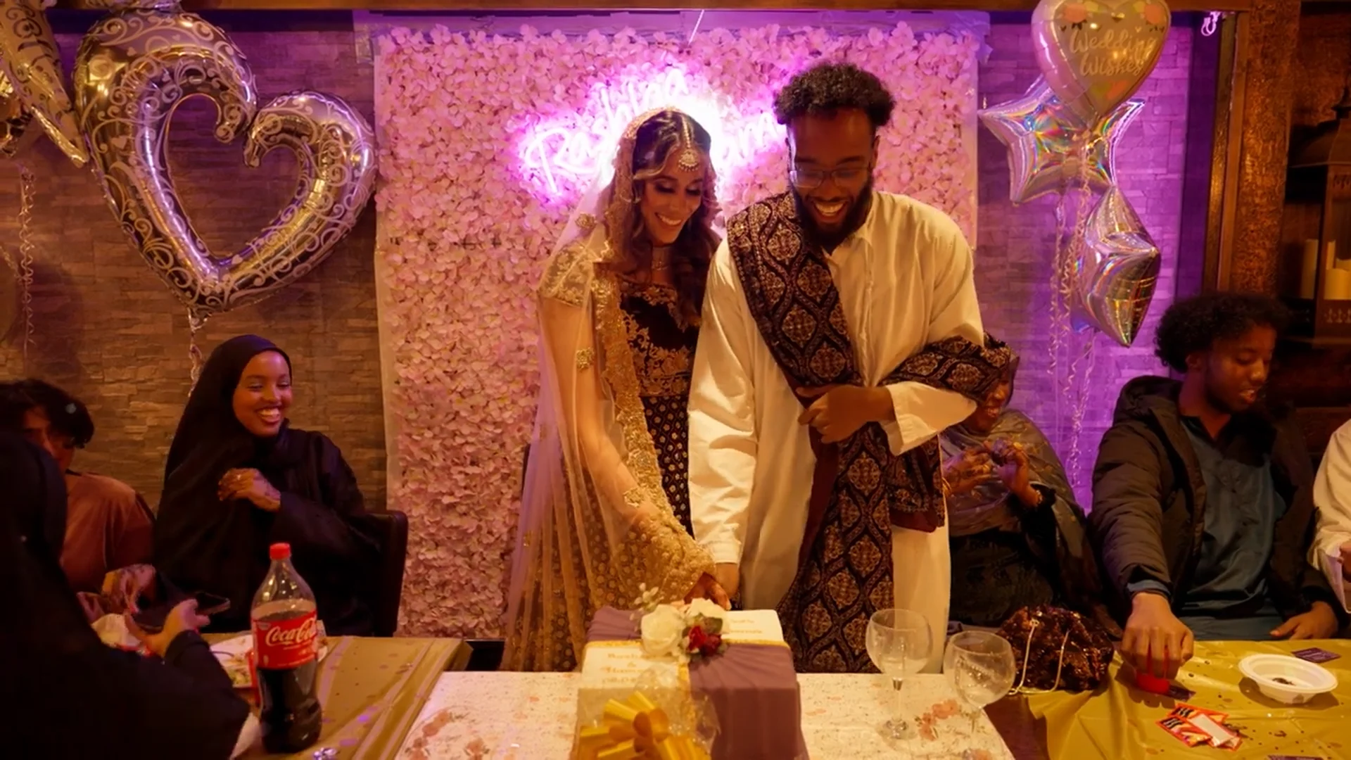 A newlywed couple laughing joyfully as they cut their wedding cake in front of a pink floral wall and neon sign, surrounded by guests at a festive reception.