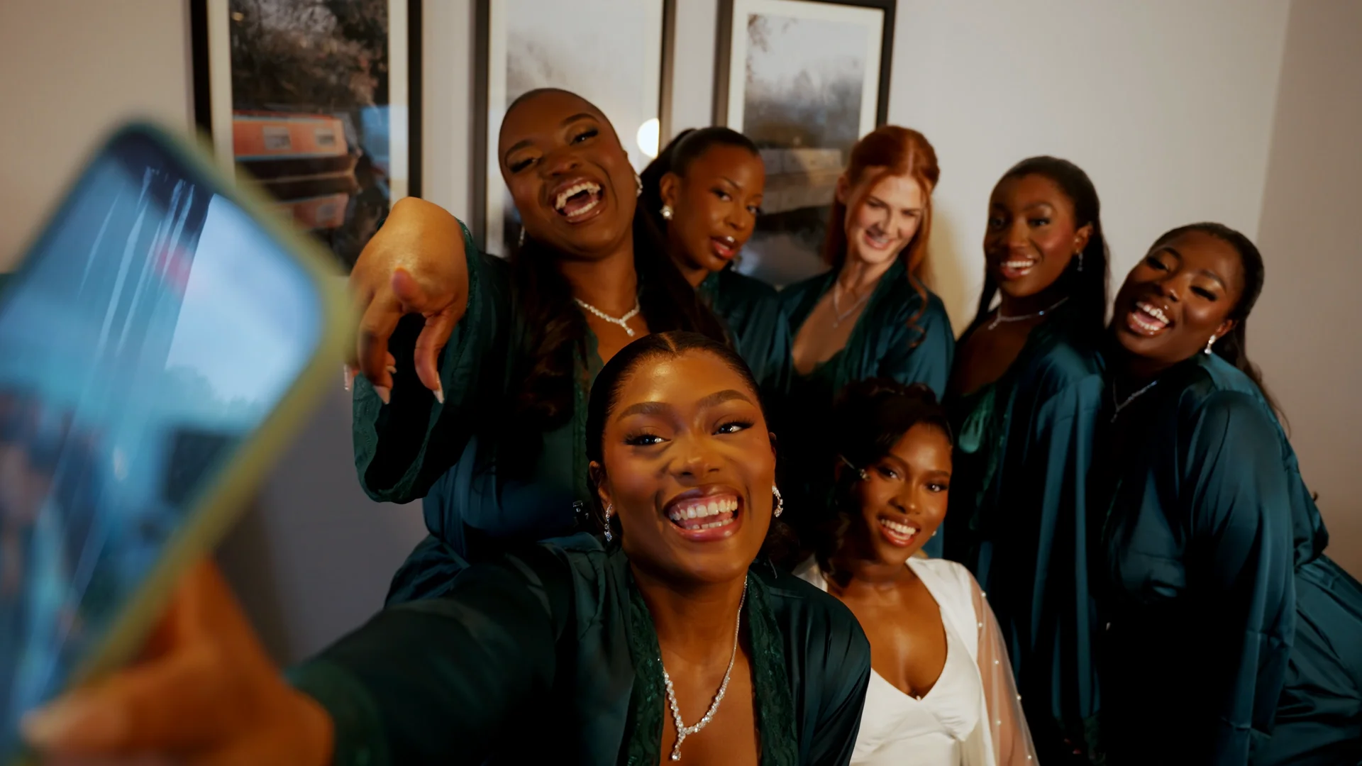 A joyful bride and her bridesmaids wearing matching green satin robes take a selfie together while laughing and smiling.