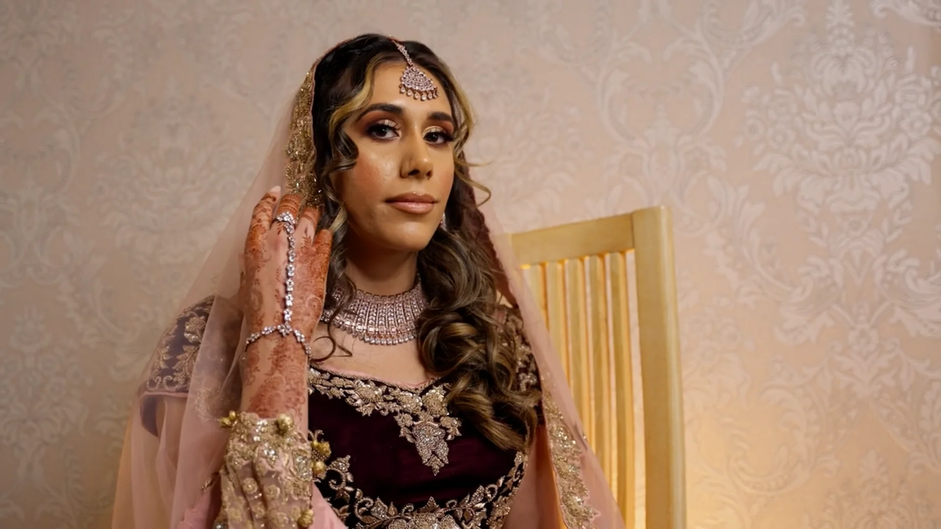 A stunning South Asian bride seated on a wooden chair, wearing a heavily embroidered dark outfit with a sheer pink veil and intricate gold jewellery.