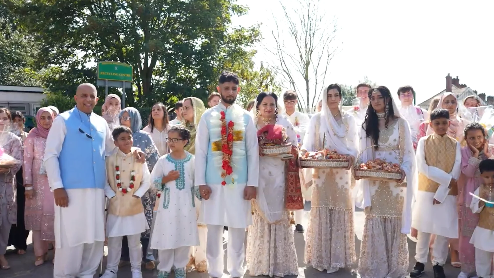 A large multi-generational wedding party in traditional South Asian attire poses outdoors in bright sunlight.