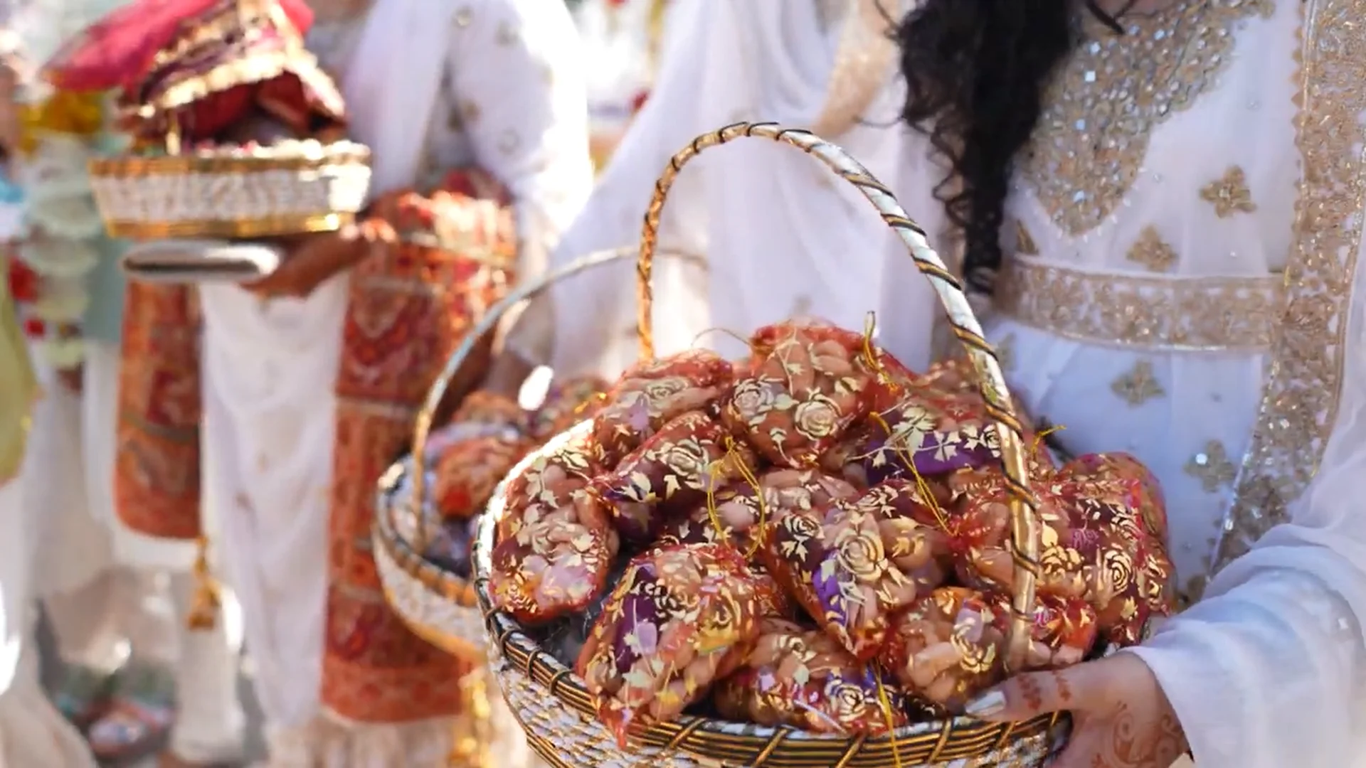 Guests dressed in ornate white and gold traditional attire hold woven baskets filled with decorative parcels during a vibrant cultural wedding celebration.