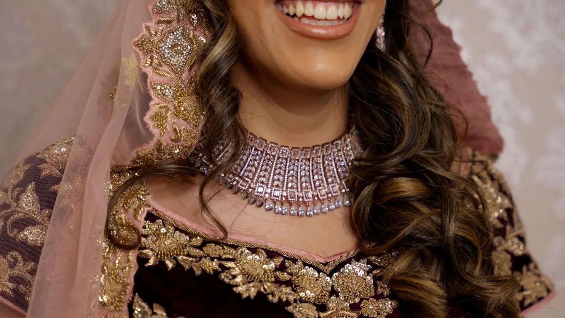 A close-up portrait of a smiling bride wearing an ornate dark velvet and gold embroidered outfit, paired with a sparkling crystal choker necklace and a sheer pink veil.