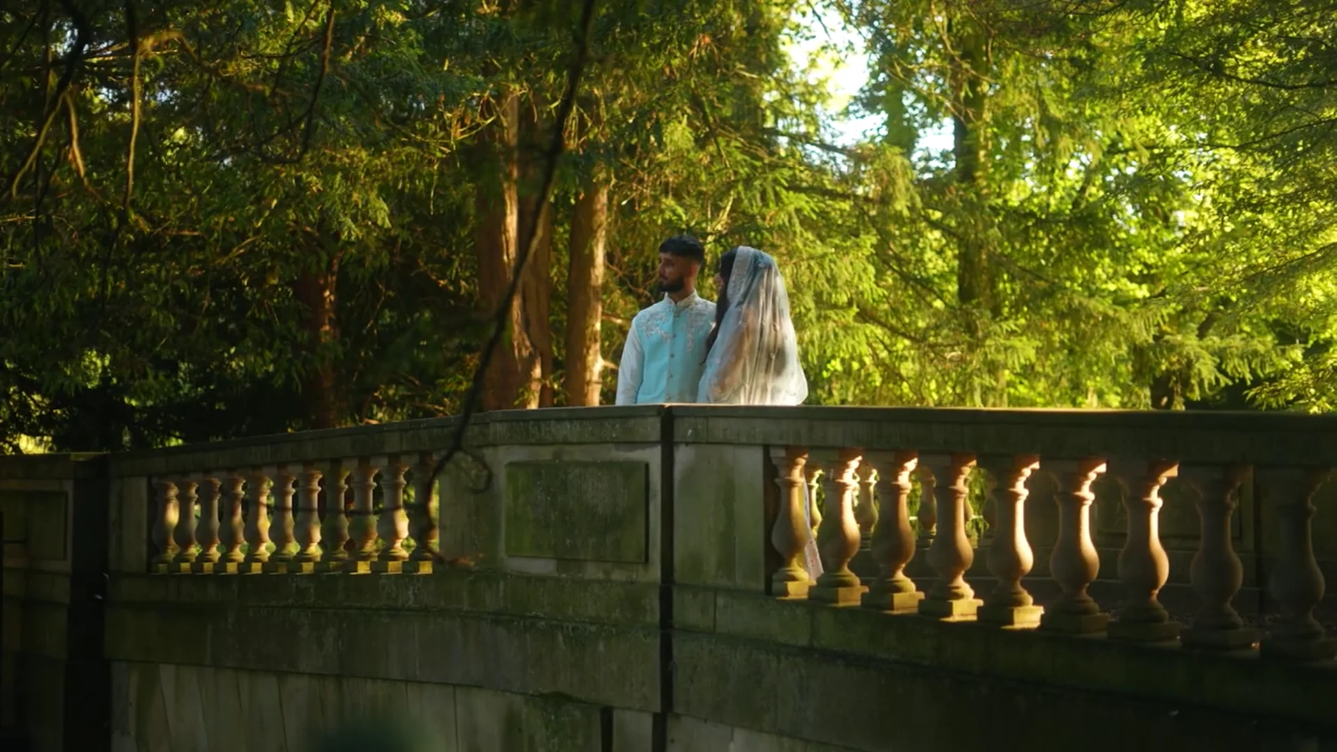 A newlywed couple stands together on a stone bridge framed by a lush woodland background, illuminated by warm sunlight filtering through the trees.