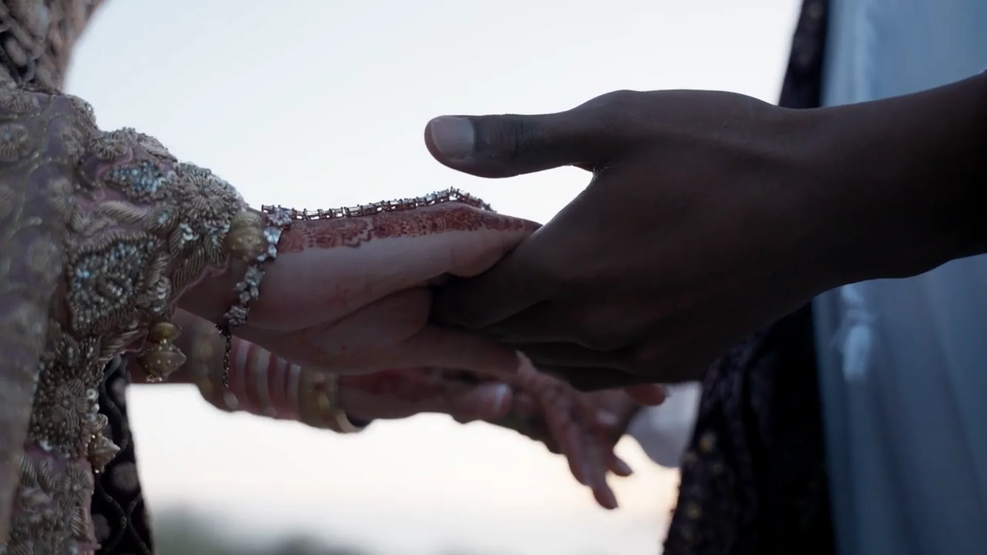 An intimate close-up shot capturing a bride and groom holding hands, highlighting intricate henna designs and ornate traditional garments against a soft, bright background.