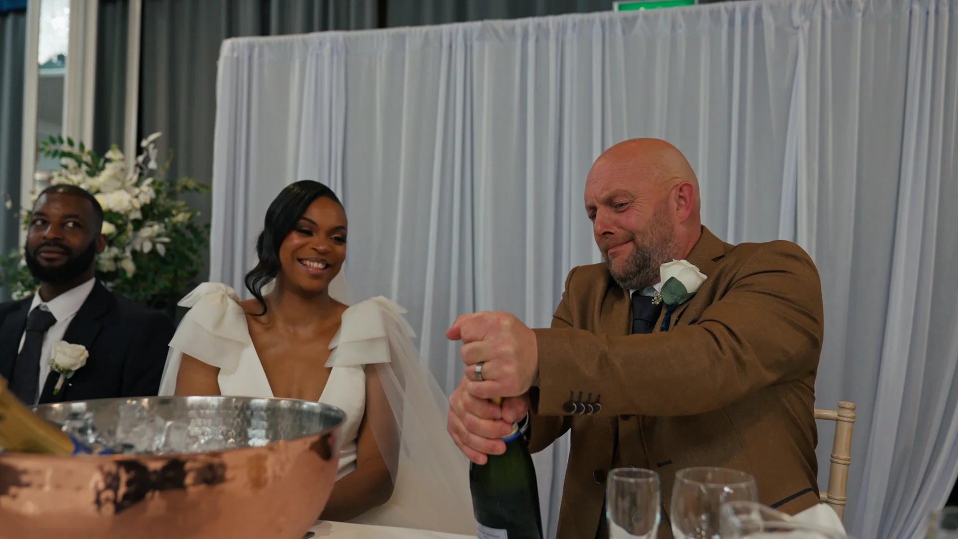 An interracial couple and a guest enjoy a joyful moment at the top table during a wedding reception as champagne is opened.