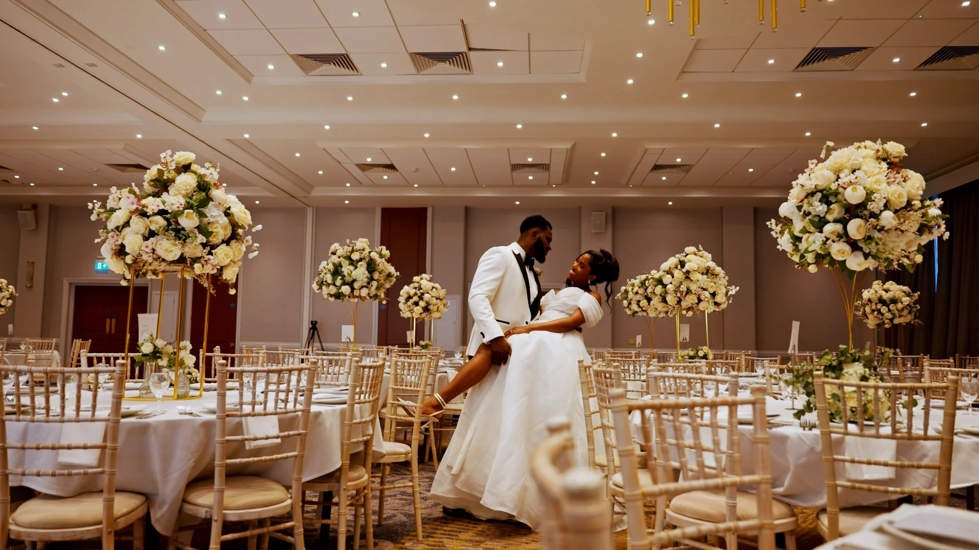 A groom in a white tuxedo lifts his bride in a white off-the-shoulder gown in the centre of a large ballroom decorated with round tables and tall floral centrepieces.
