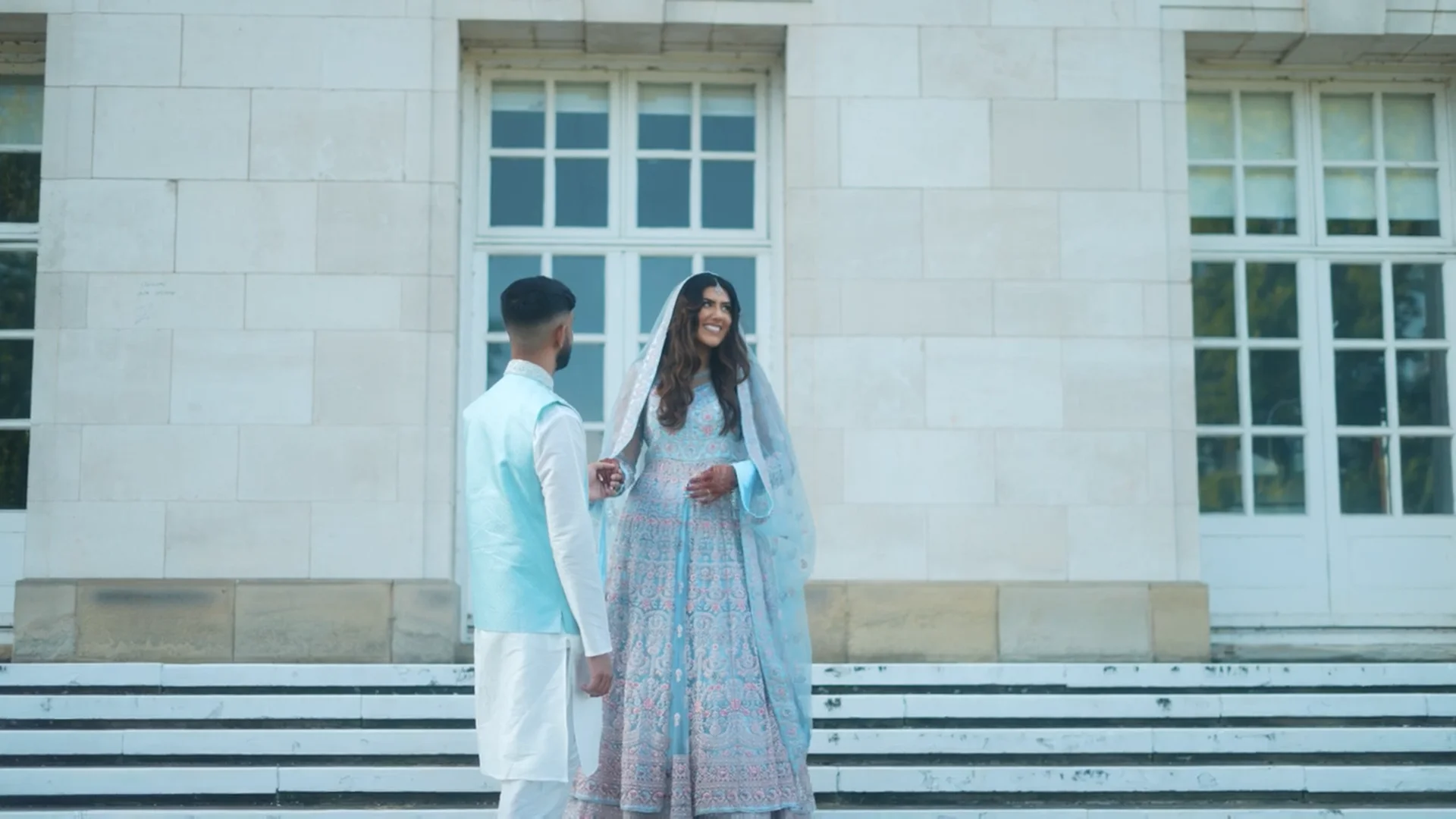 A newlywed couple in elegant pastel blue South Asian attire stand outside a grand stone building, holding hands and smiling at one another.