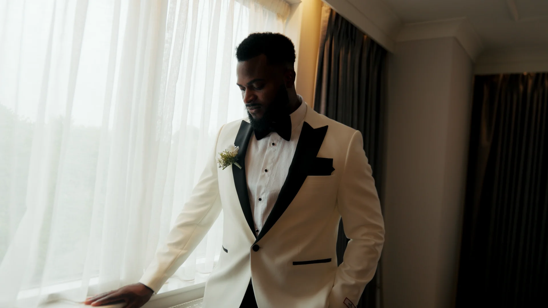 A handsome groom in a white tuxedo jacket and black bow tie stands by a bright window, looking thoughtfully downwards during his wedding preparations.