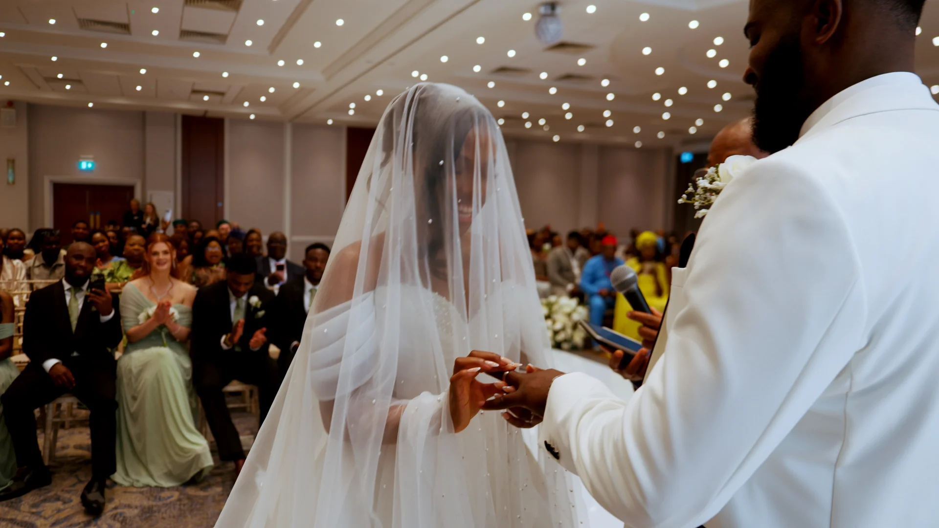 A smiling bride wearing an off-the-shoulder gown and a pearl-embellished veil exchanges rings with her groom during an indoor wedding ceremony.