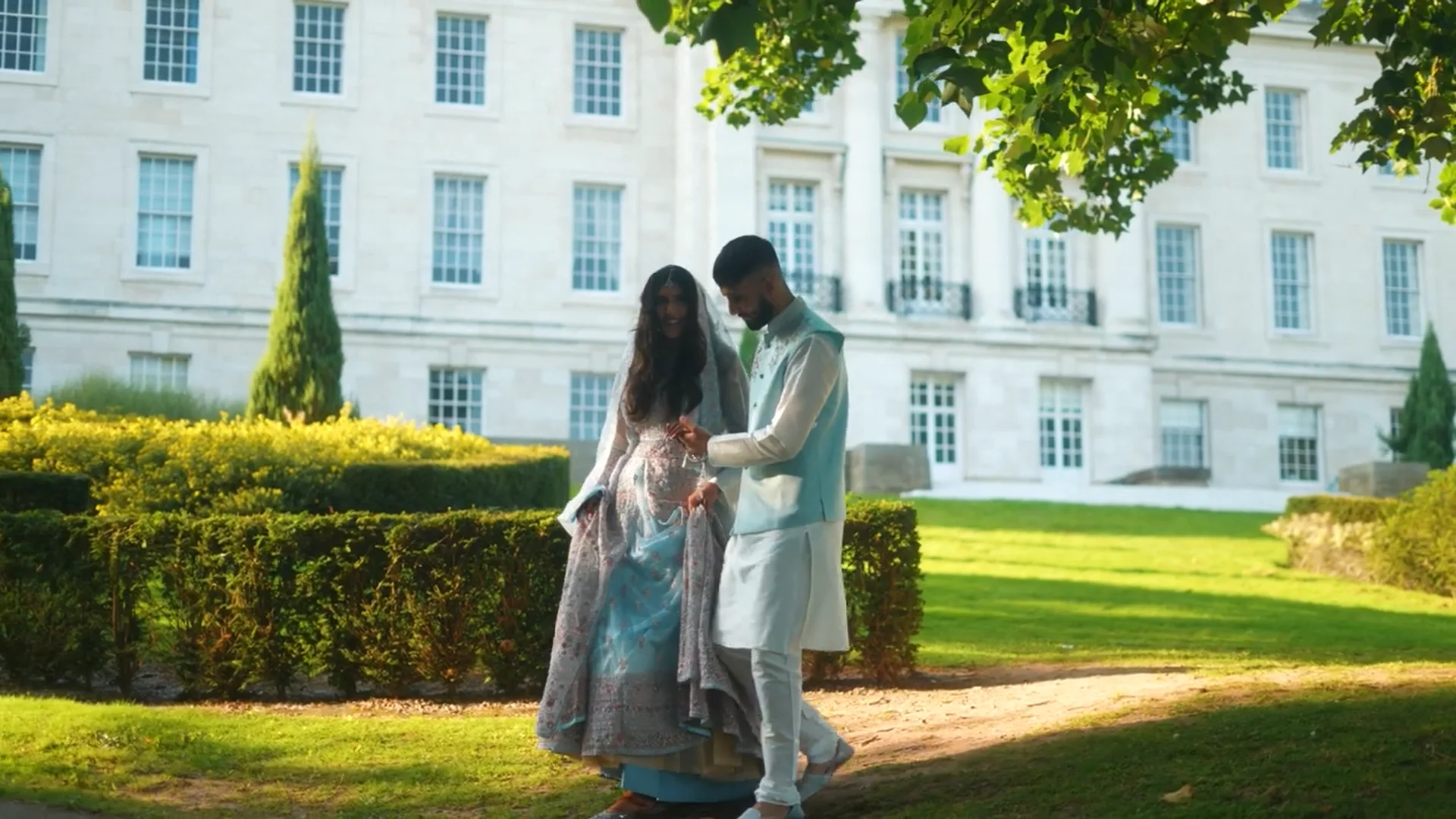 A newlywed couple walks hand in hand through the manicured gardens of a grand country house on a sunny wedding day.