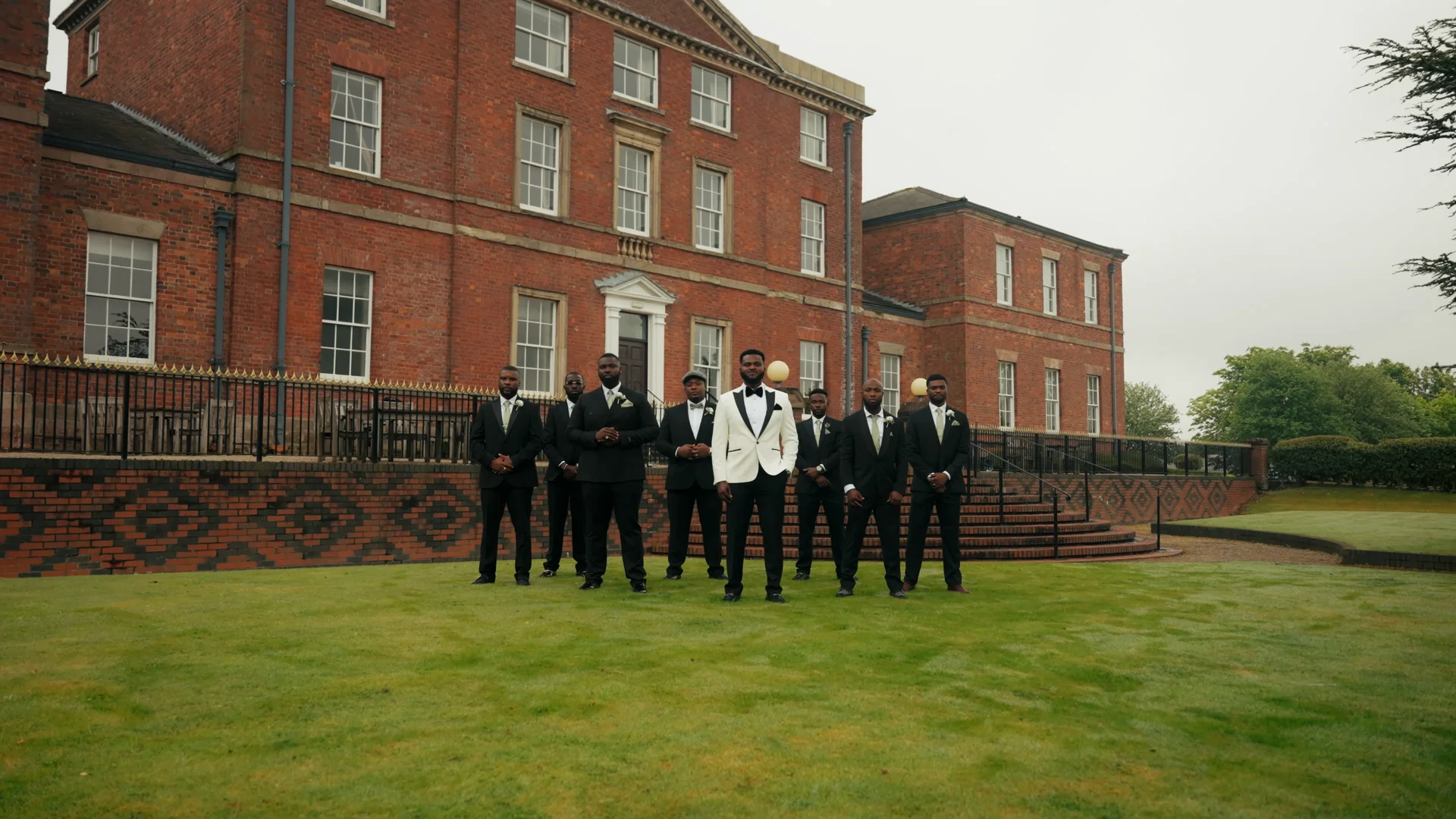 A stylish groom in a white tuxedo jacket and his groomsmen in classic black tuxedos pose confidently on the lawn in front of a grand brick manor house.