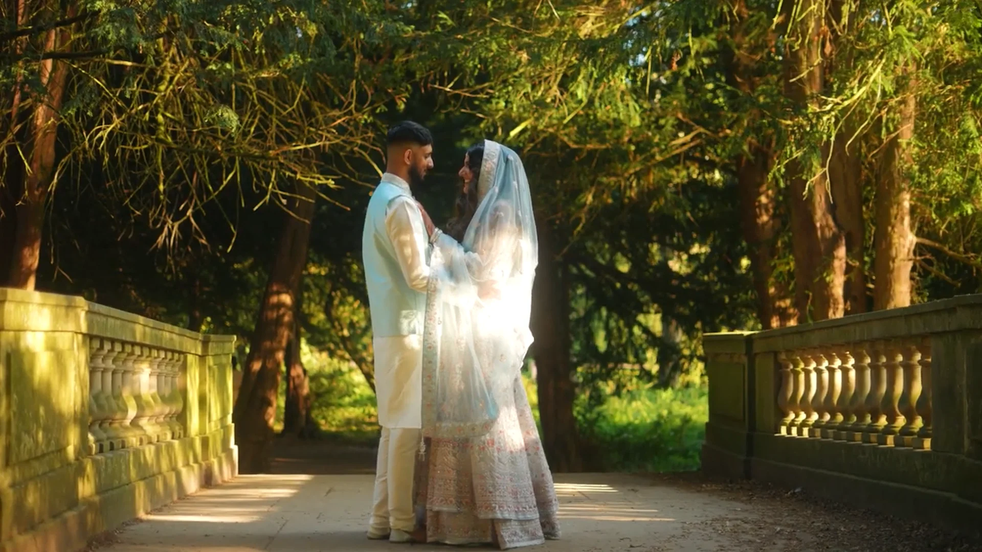 A newlywed couple embraces on a stone bridge surrounded by a sunlit woodland, with dappled light filtering through the trees.