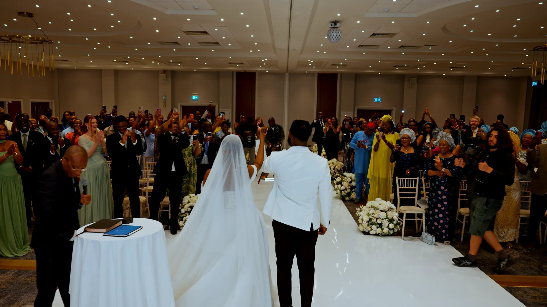 Newlyweds stand hand-in-hand facing an applauding crowd of wedding guests inside a spacious, modern hotel ballroom.