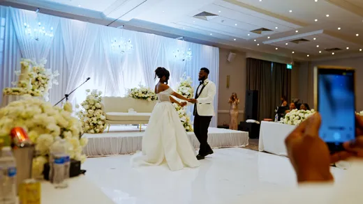 A newlywed couple shares their first dance on a white gloss dance floor in a brightly lit ballroom decorated with white floral arrangements and draping.
