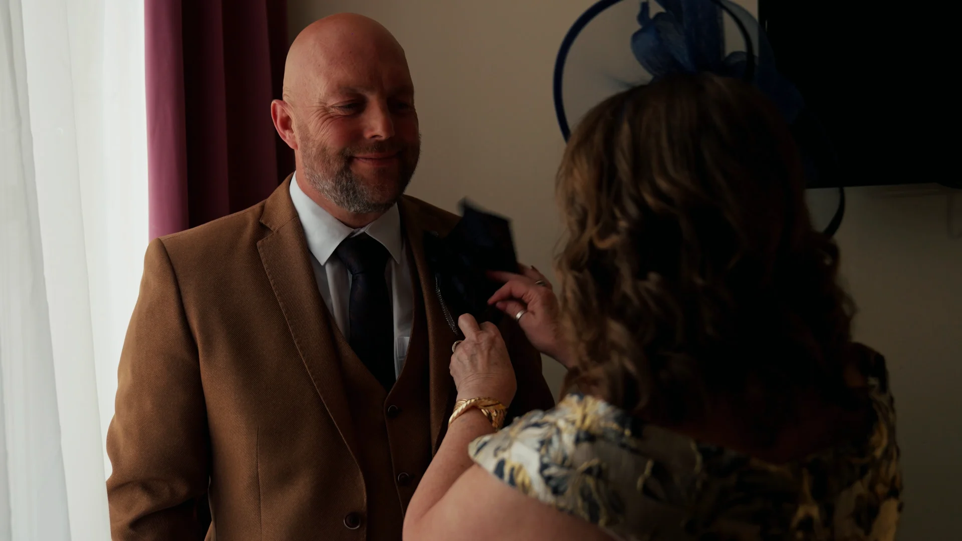 A smiling groom wearing a tan three piece suit gets the finishing touches on his buttonhole from a guest wearing a fascinator on the wedding morning.