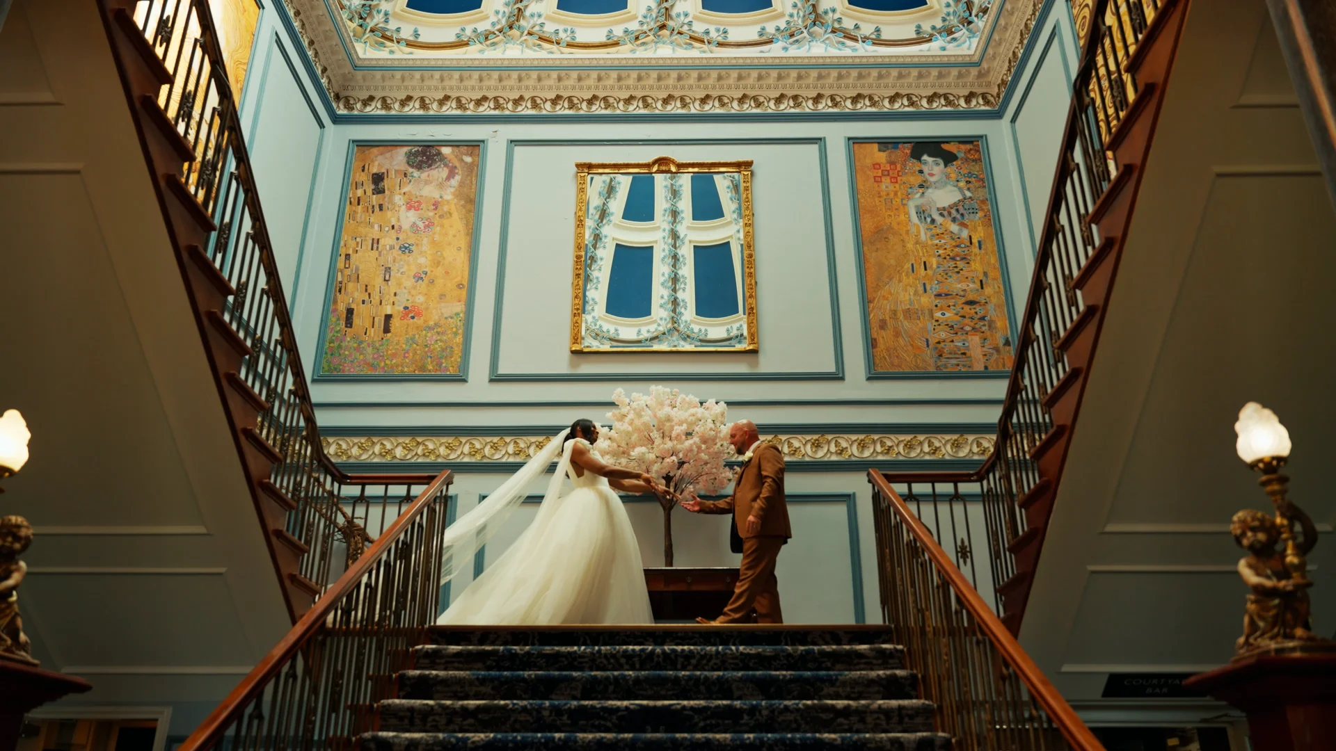An interracial newlywed couple stands on a grand carpeted staircase inside a lavish country house venue beneath high ceilings and large decorative artwork.