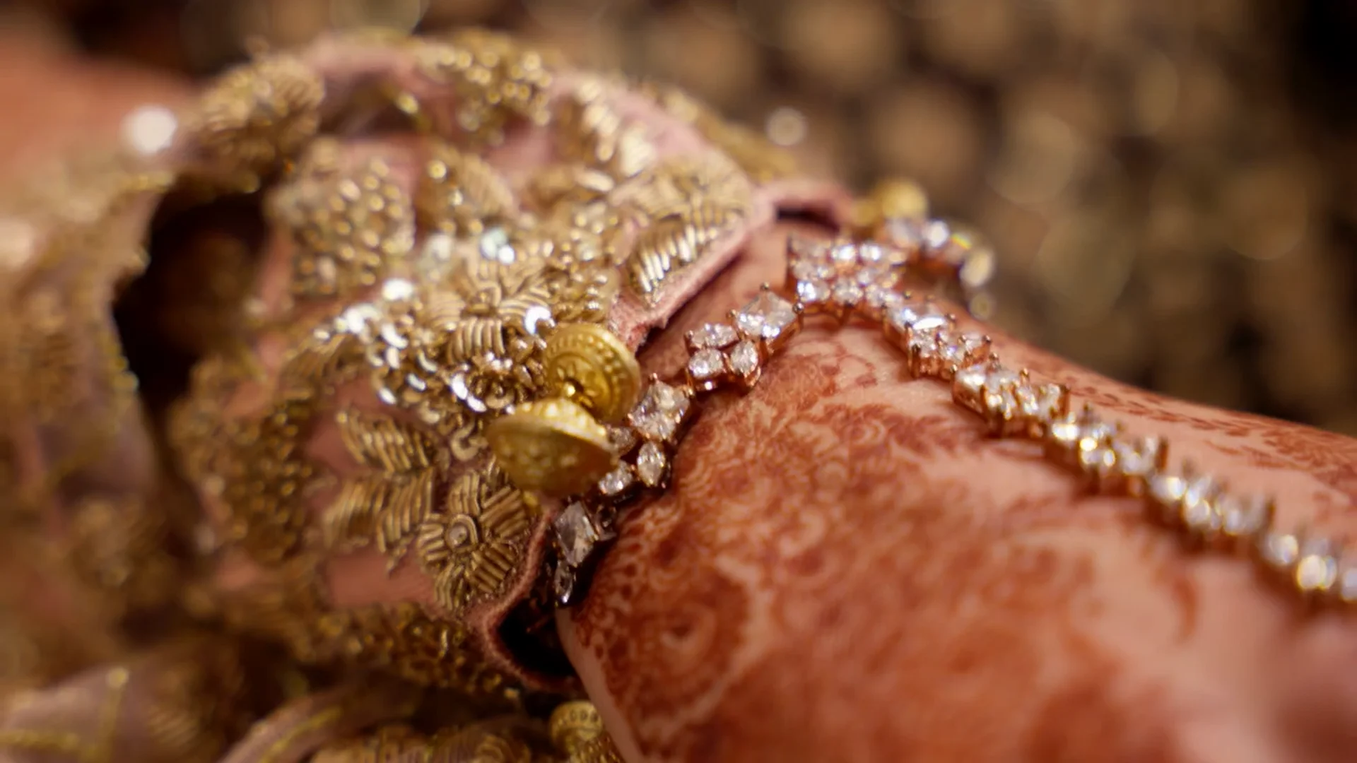 An extreme close-up highlights intricate henna patterns on a bride's hand adorned with a delicate diamond bracelet and heavily embroidered gold garments.