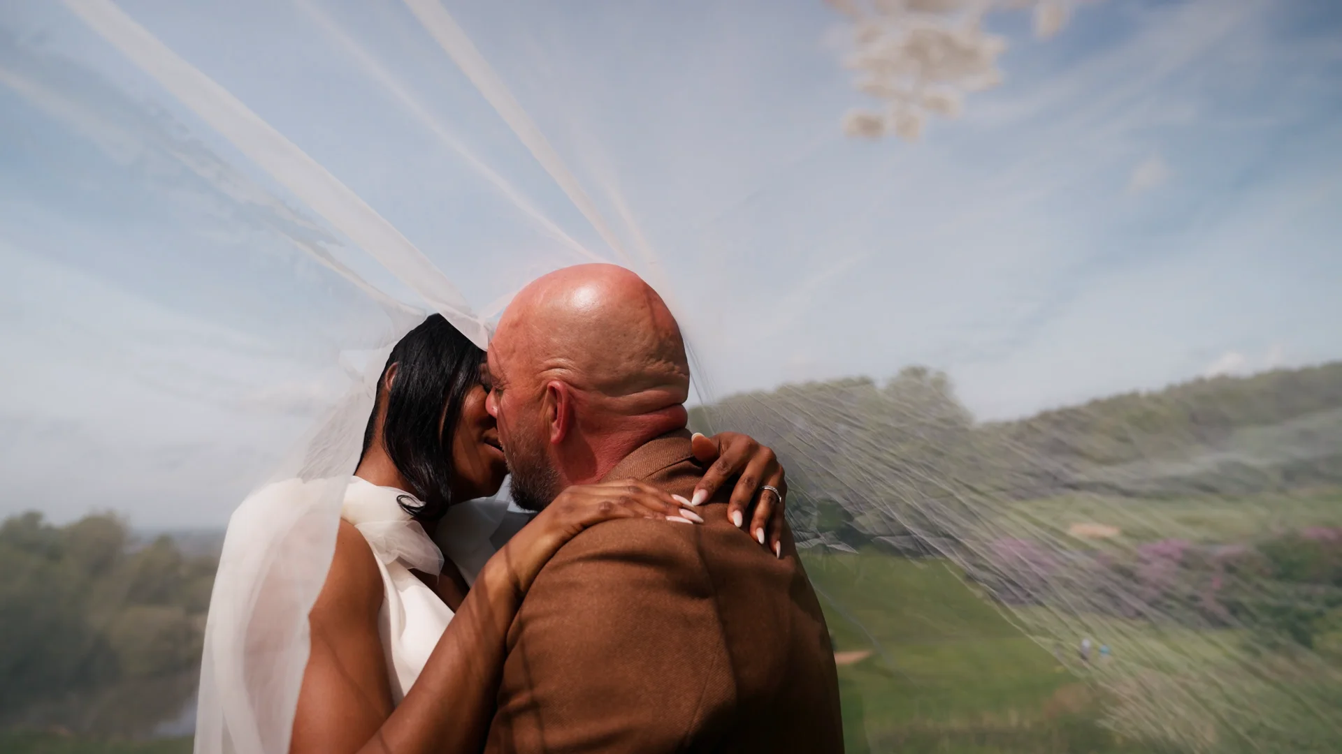 An interracial newlywed couple share a tender kiss beneath a sheer white veil outdoors against a scenic landscape backdrop.