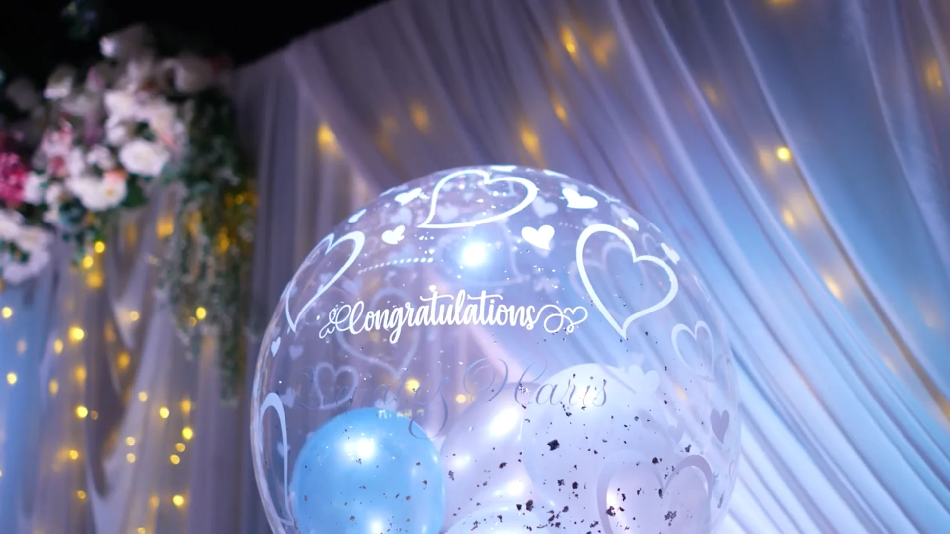 A clear decorative wedding balloon featuring white printed hearts and the word congratulations sits in front of draped fabric and fairy lights.