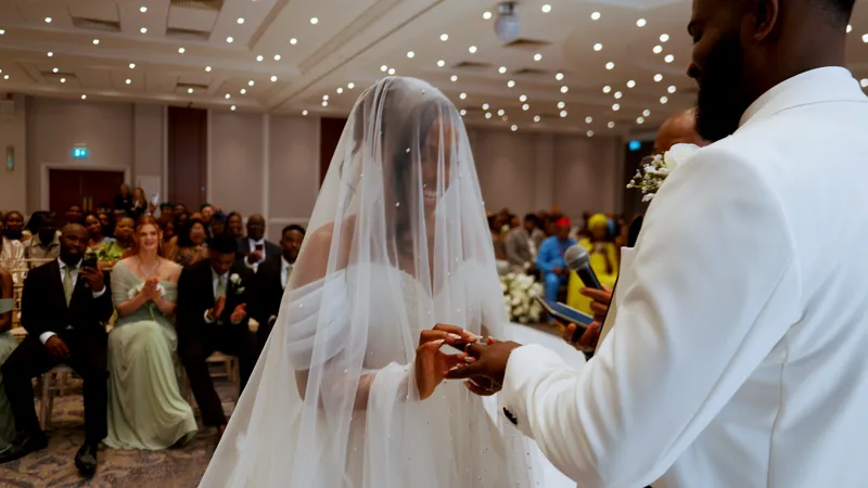 A Black bride wearing a pearl-embellished veil and off-the-shoulder gown exchanges rings with her groom in a white tuxedo during an indoor wedding ceremony.