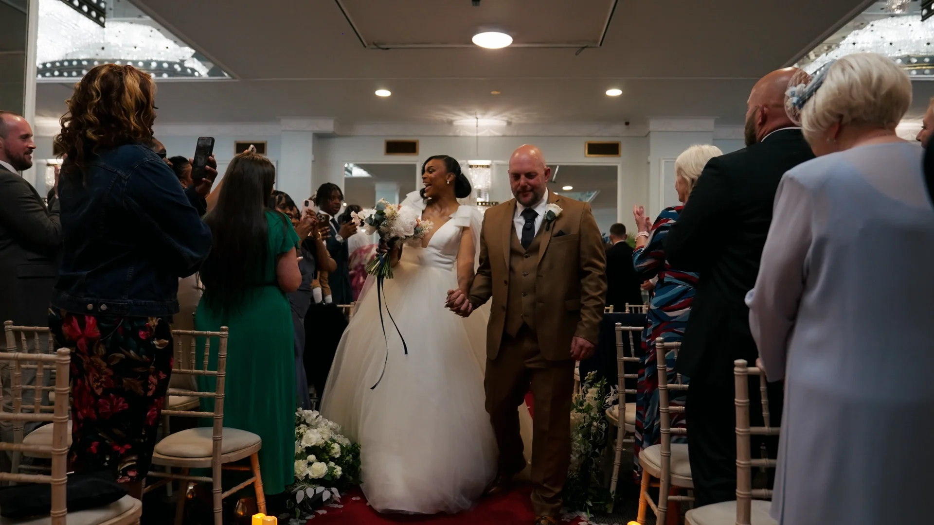 Newlyweds joyfully walk hand in hand down the aisle amidst cheering guests after their hotel wedding ceremony.