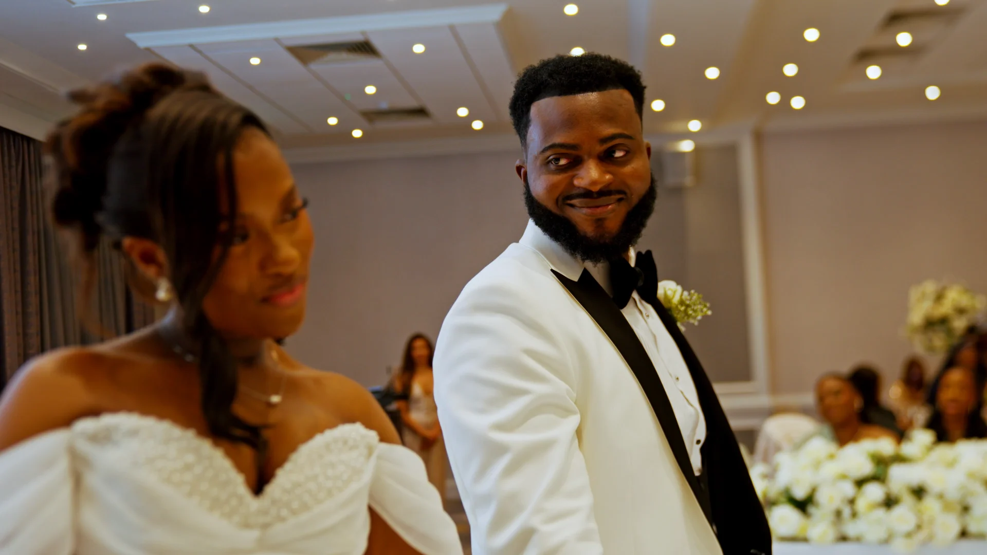 A newlywed couple shares a stolen glance during their wedding reception, with the groom wearing a white tuxedo jacket and the bride in an off-the-shoulder gown.