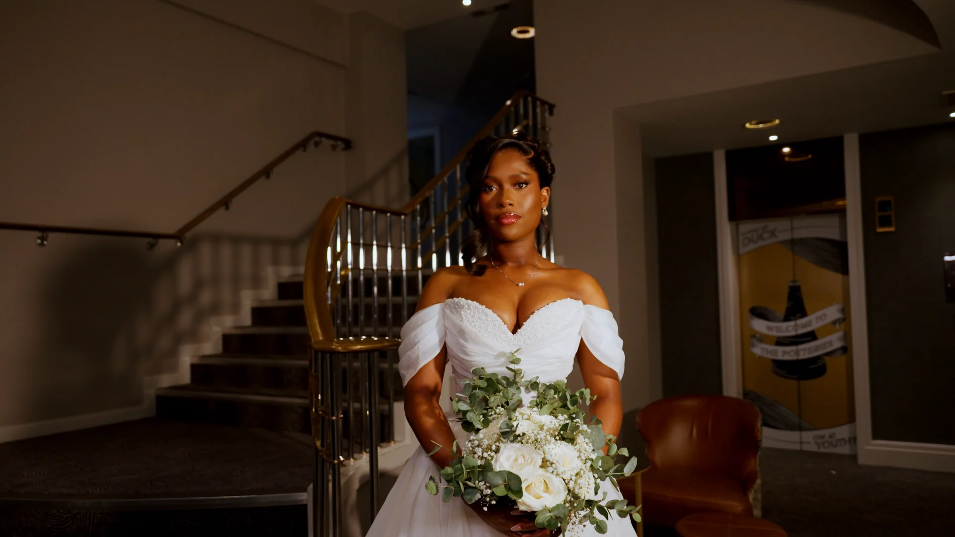 A stunning black bride in an off-the-shoulder white gown holds a lush rose and eucalyptus bouquet while posing elegantly indoors near a grand staircase.