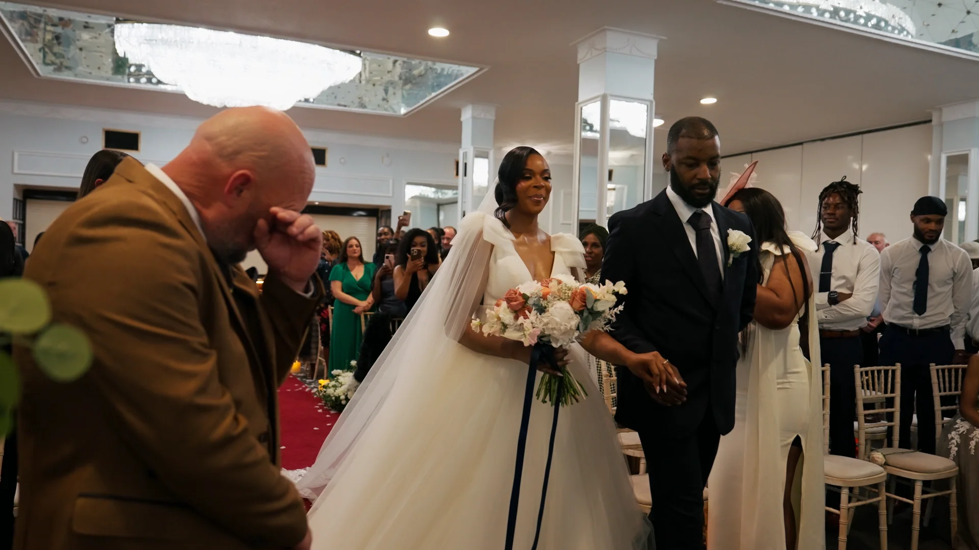 A newlywed bride and groom walk down the aisle together while a guest in the foreground wipes away emotional tears of joy.