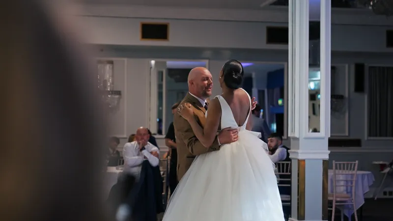 A newlywed couple shares their first dance in an elegant ballroom, with the groom wearing a brown suit and the bride in a classic white gown with a voluminous tulle skirt.