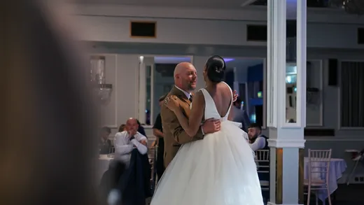A newlywed couple shares their first dance in an elegant ballroom, with the groom wearing a brown suit and the bride in a classic white gown with a voluminous tulle skirt.