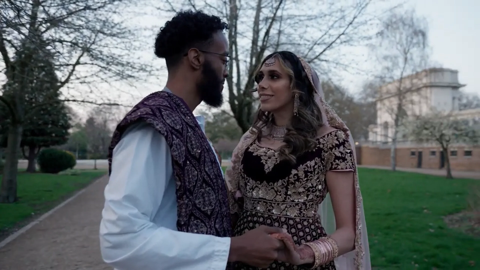 A newlywed couple shares a tender glance outdoors in a park setting, wearing traditional embroidered garments and holding hands.