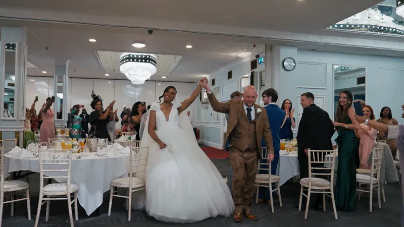 Newlyweds enter the wedding reception hall hand in hand, smiling brightly as guests seated at round tables applaud and capture the moment on their phones.