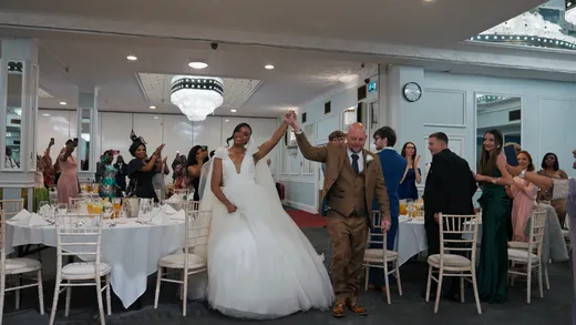 Newlyweds enter the wedding reception hall hand in hand, smiling brightly as guests seated at round tables applaud and capture the moment on their phones.