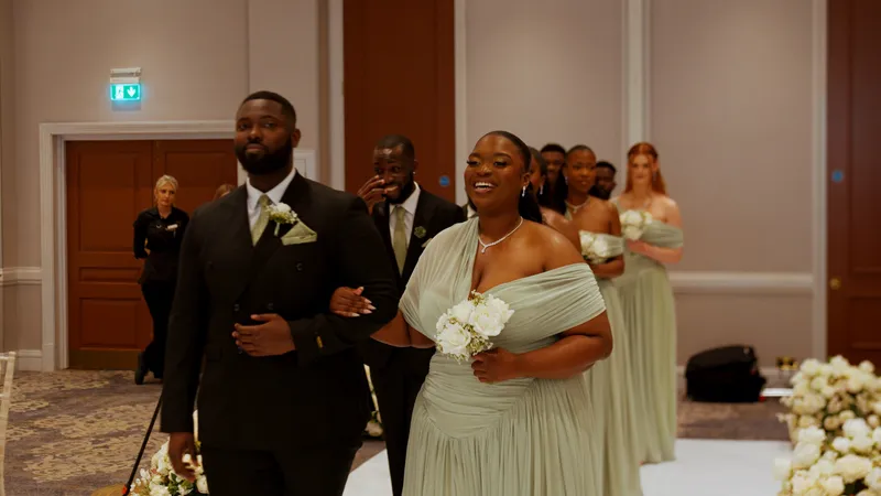 A smiling bridesmaid in a sage green off-the-shoulder gown walks down the aisle arm in arm with a groomsman in a black suit, followed by a wedding party.