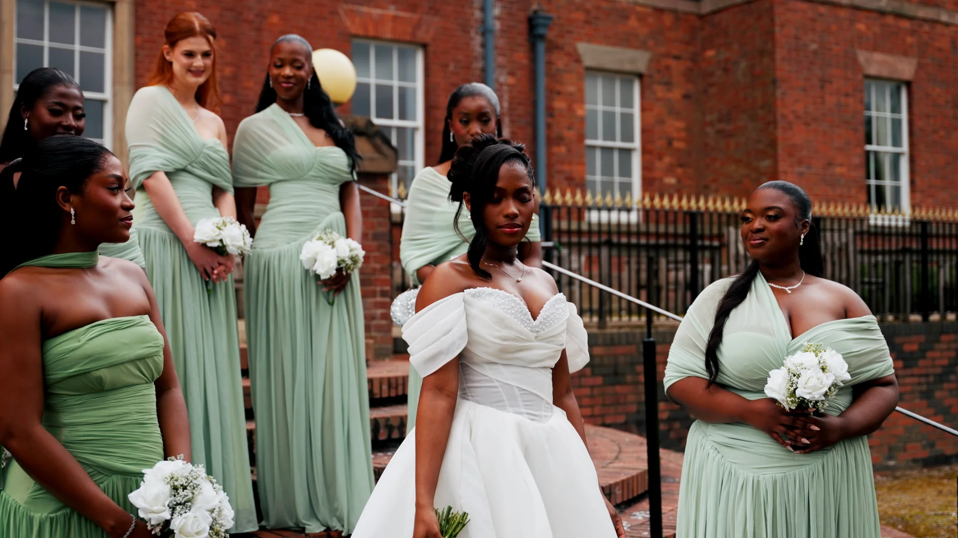 A stunning bride in an off-the-shoulder white ballgown stands proudly surrounded by her bridal party wearing matching sage green dresses outside a red brick building.