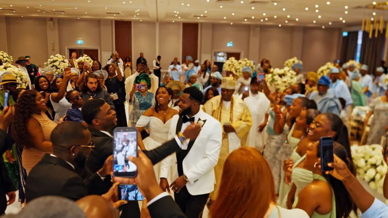 A joyous newlywed couple in a white tuxedo and off-the-shoulder gown are surrounded by cheering wedding guests capturing the grand reception entrance on their phones.