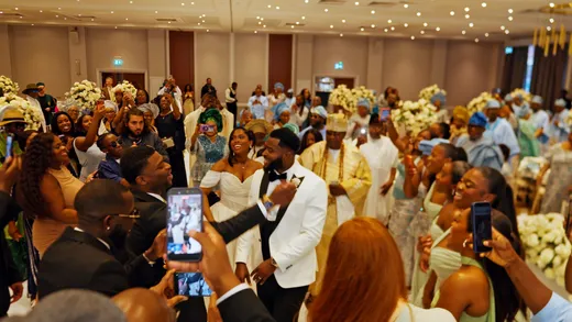 A joyous newlywed couple in a white tuxedo and off-the-shoulder gown are surrounded by cheering wedding guests capturing the grand reception entrance on their phones.