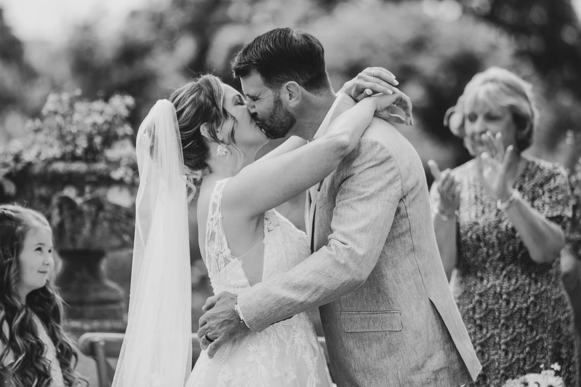 A newlywed couple share their first kiss outdoors while family members applaud and watch on with joy.