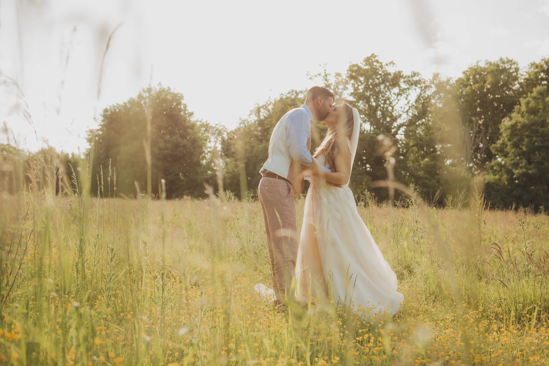 A newlywed couple shares a tender kiss bathed in warm golden hour sunlight amidst a lush wildflower meadow.