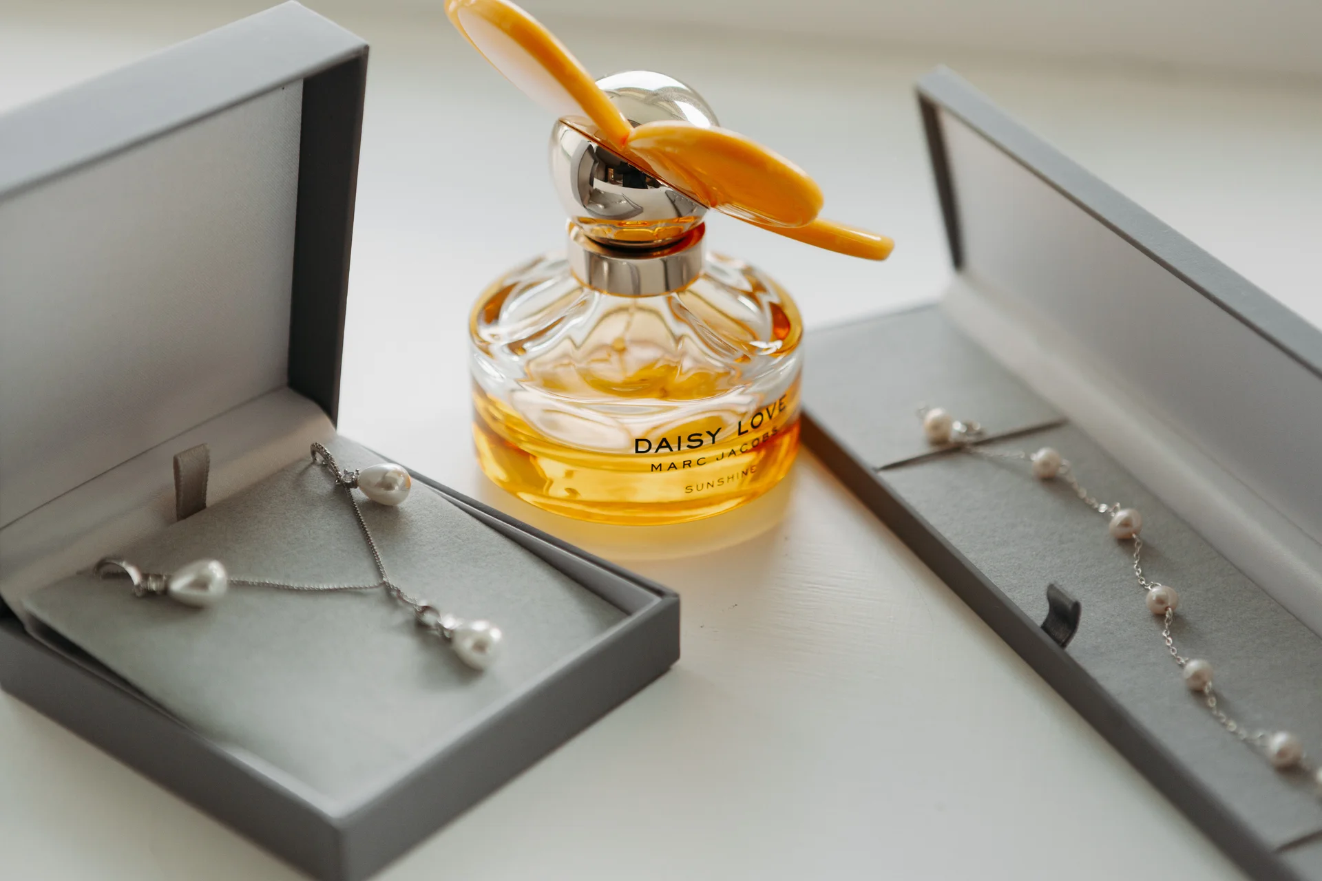 An elegant bridal flat lay features a bottle of perfume flanked by open grey boxes displaying pearl jewellery in soft natural light.