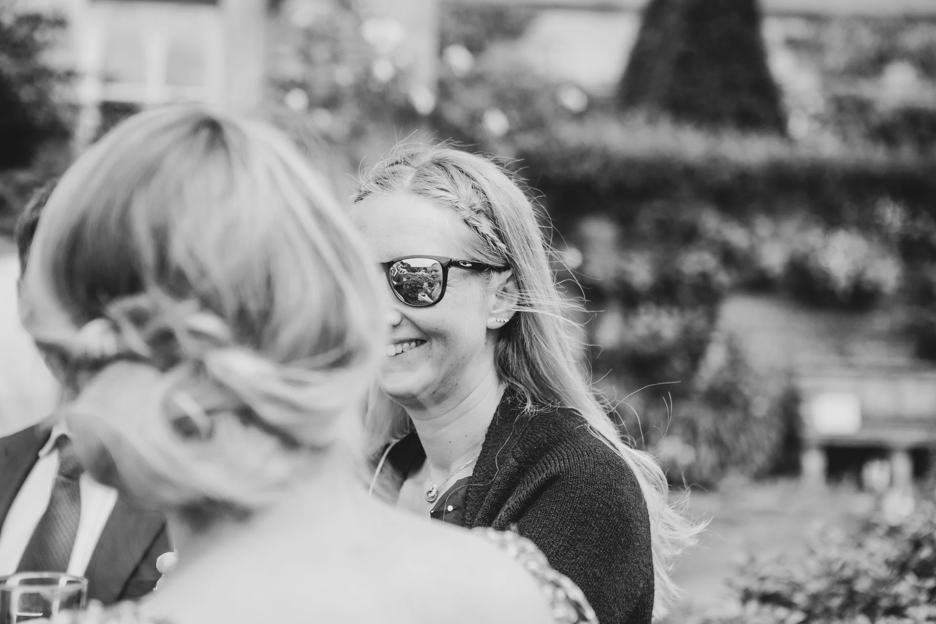 A candid black and white wedding guest shot captured from over the shoulder, showing a smiling woman wearing sunglasses with a reflection.