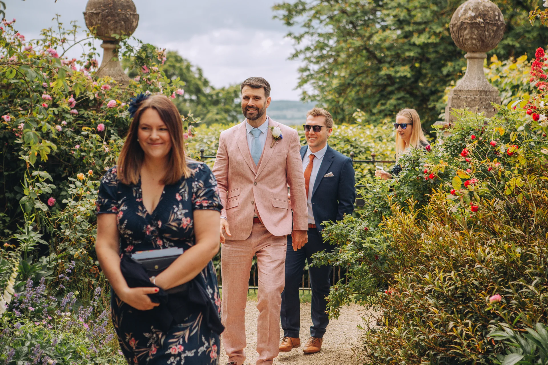 Guests and groomsmen in suits walk along a gravel path through lush manicured gardens during an outdoor wedding celebration.