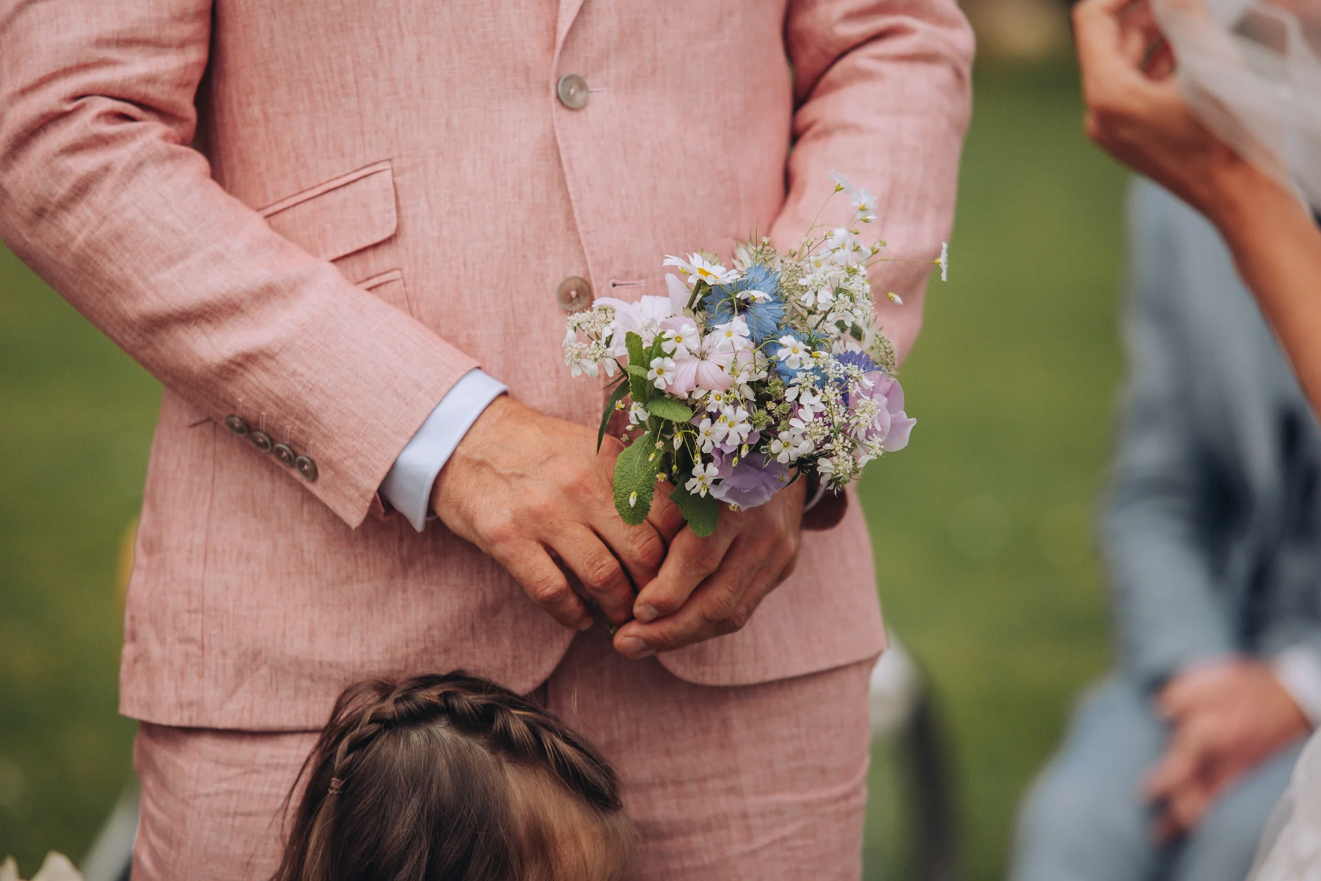 A groom wearing a pink linen suit holds a delicate wildflower meadow bouquet during an outdoor summer wedding ceremony.