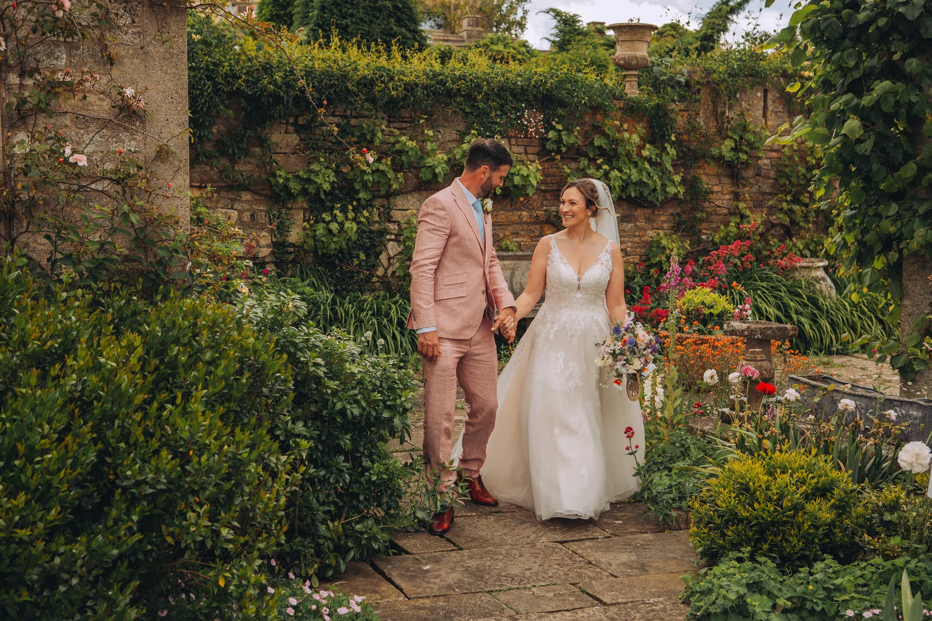 A newlywed couple smiling at each other while walking hand in hand along a stone path in a lush walled garden.