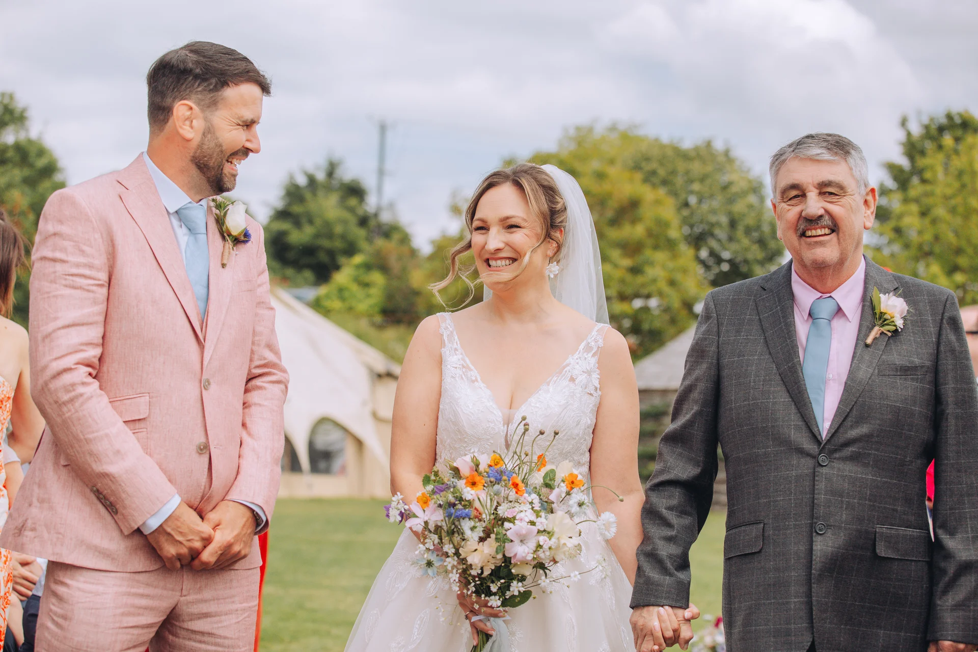 A smiling bride holding a wildflower bouquet walks down the aisle accompanied by her father, while the groom laughs happily in a pink suit.
