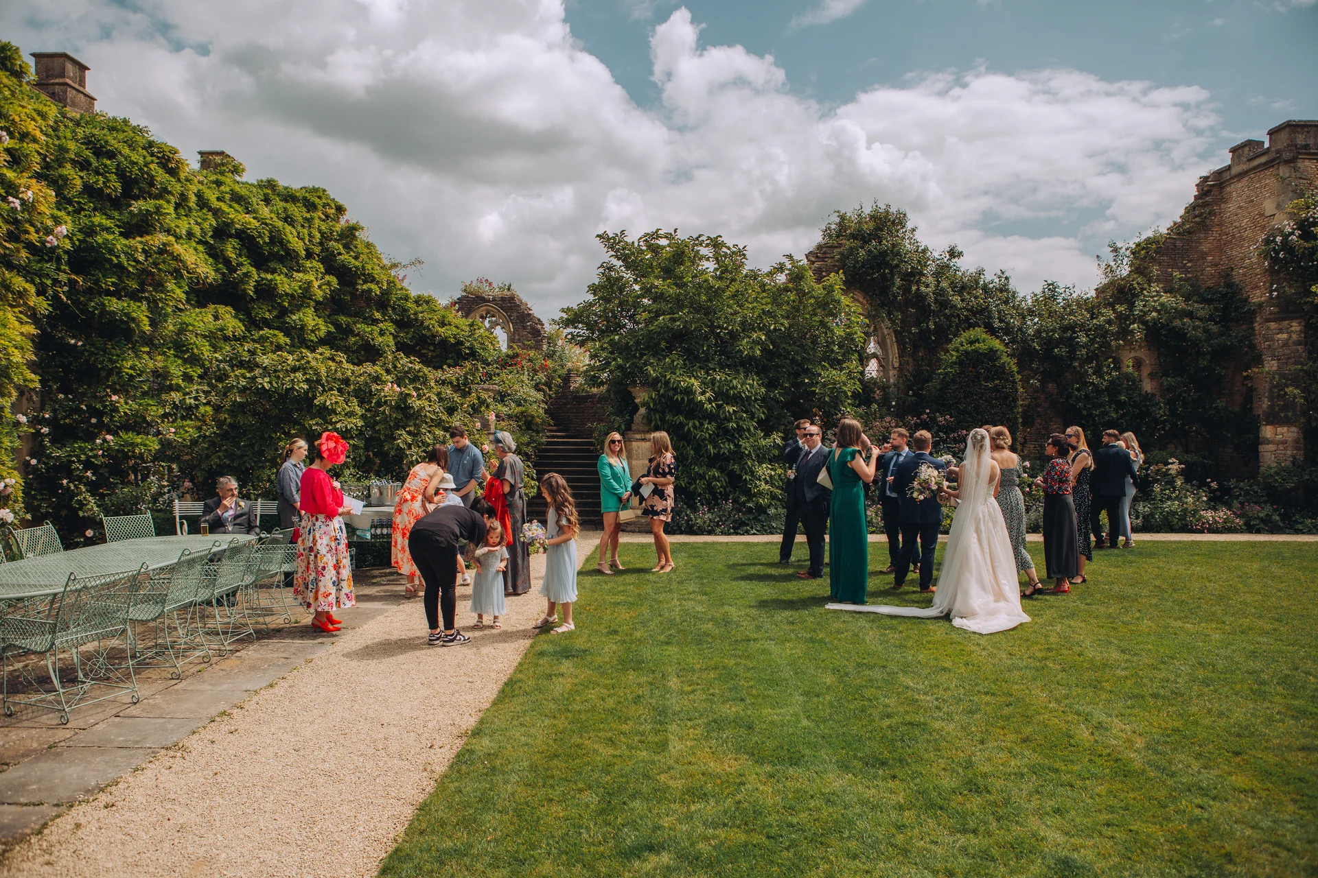 Guests mingle on a manicured lawn during an outdoor drinks reception against the backdrop of historic stone ruins and lush ivy-covered walls on a bright summer day.