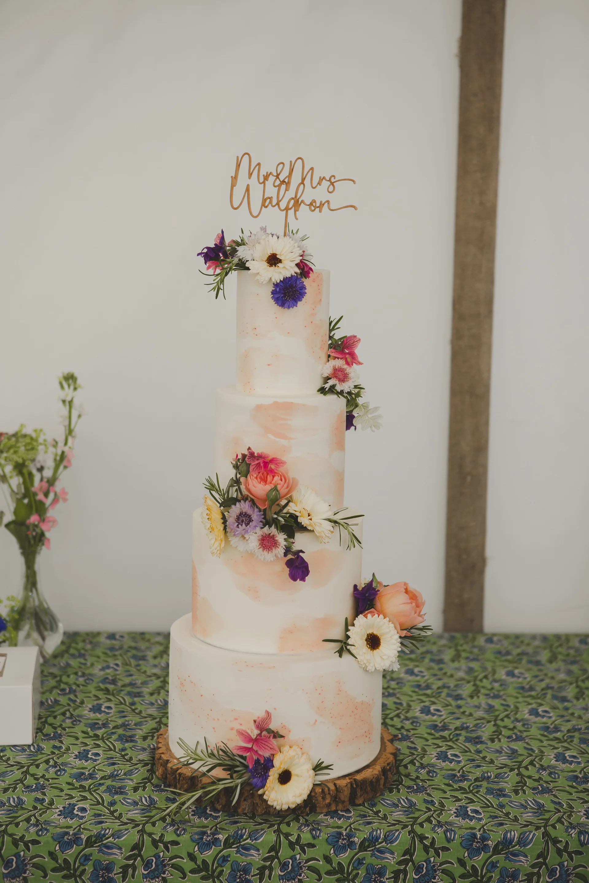 A stunning four-tier wedding cake decorated with watercolor brushstrokes and fresh colorful wildflowers sits on a wooden slice cake stand atop a green patterned tablecloth.