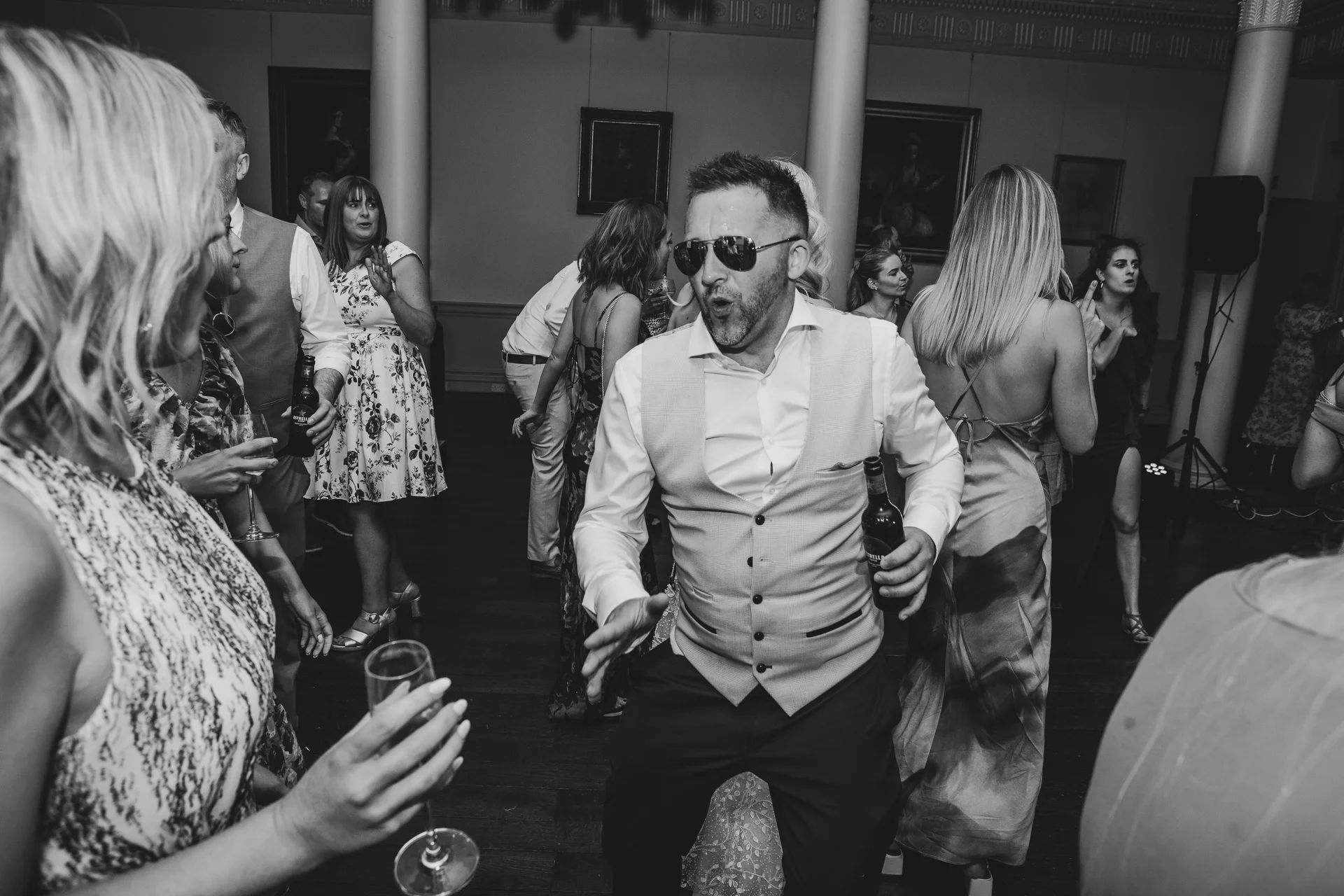 A groom wearing sunglasses and a waistcoat dances energetically on a crowded wedding reception dance floor while holding a drink.